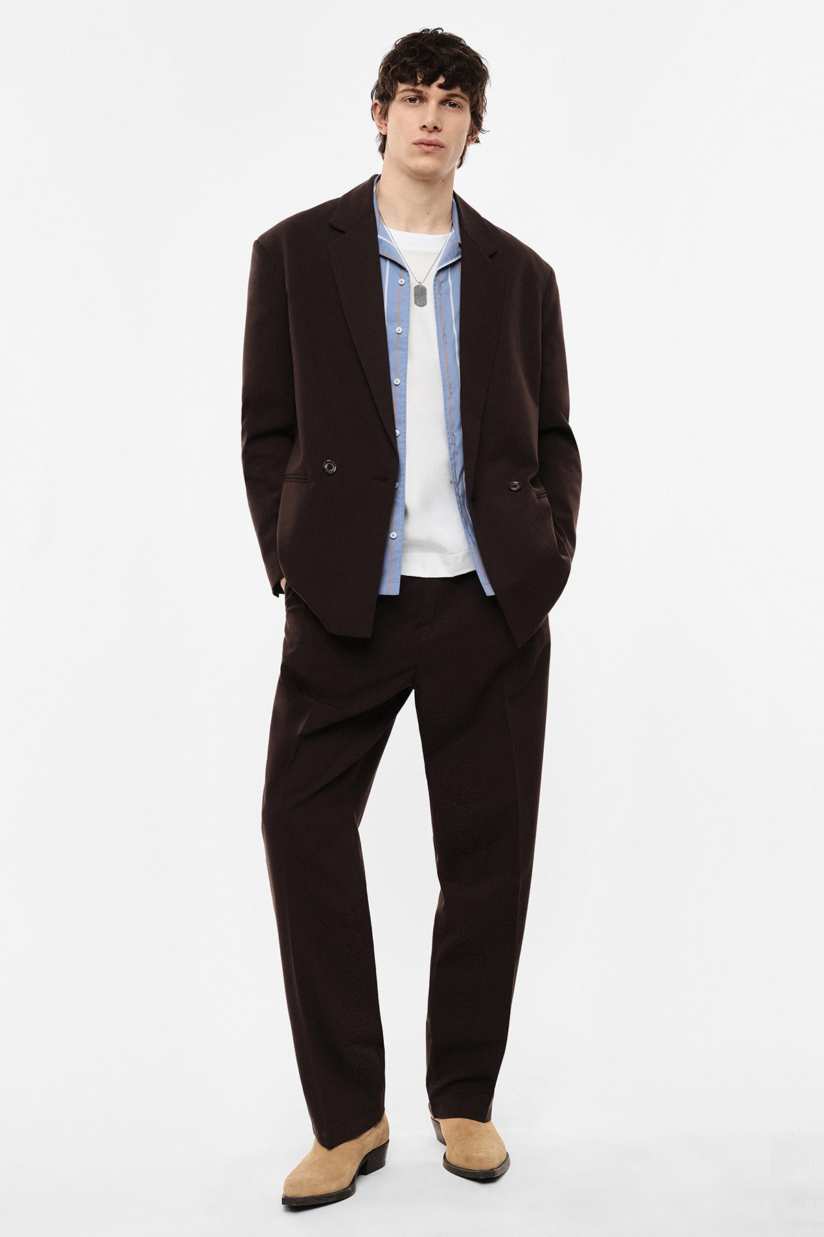 Male model in dark brown relaxed suit with light blue shirt over white tee and beige suede shoes on white background