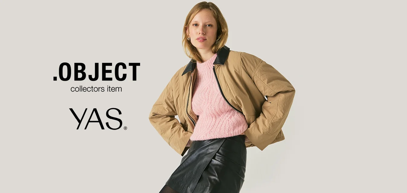 Giovane donna in posa con le mani sui fianchi, che indossa una giacca color cammello, un maglione rosa, una gonna di pelle nera e calze nere, seduta su una sedia davanti a uno sfondo neutro.