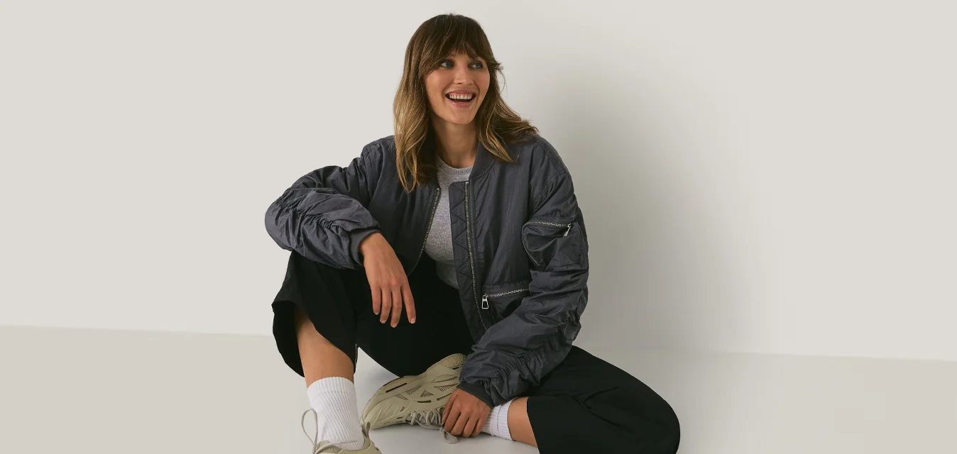 Giovane donna in abbigliamento casual e sneakers seduta sul pavimento contro un muro bianco semplice, sorridente e che guarda di lato.