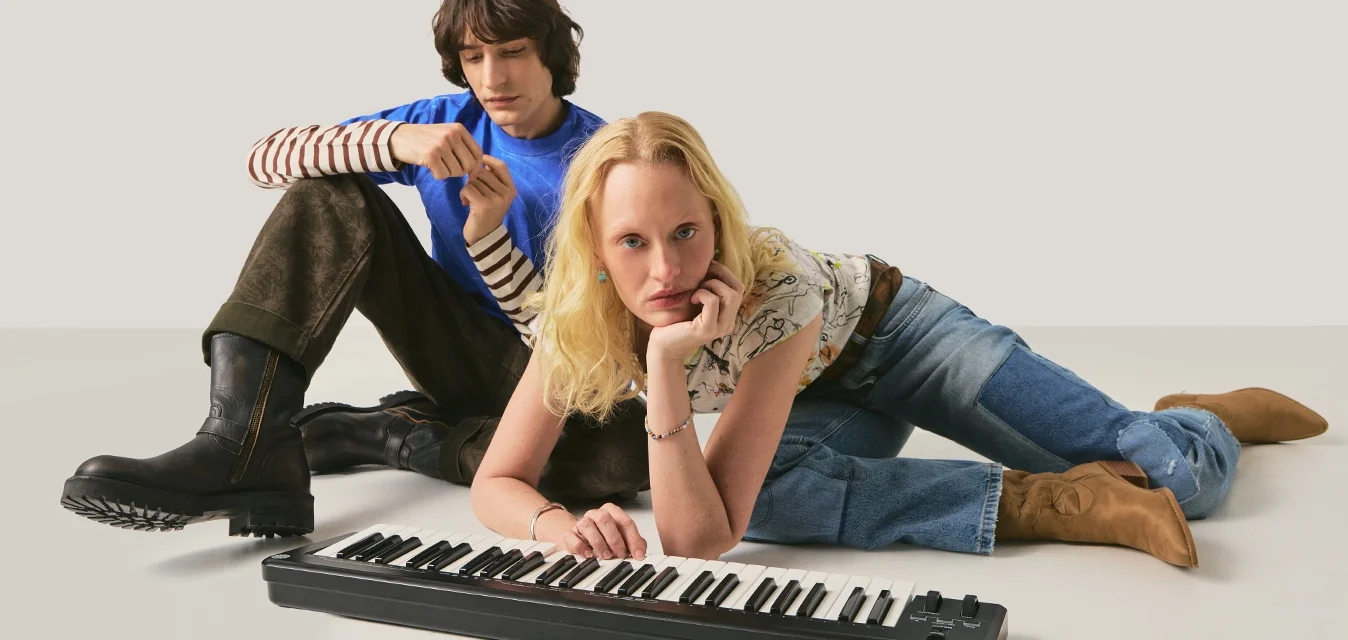 Due giovani seduti a terra con una tastiera elettronica; uno si appoggia alla tastiera, l'altro è seduto dietro con stivali e maglietta a righe.