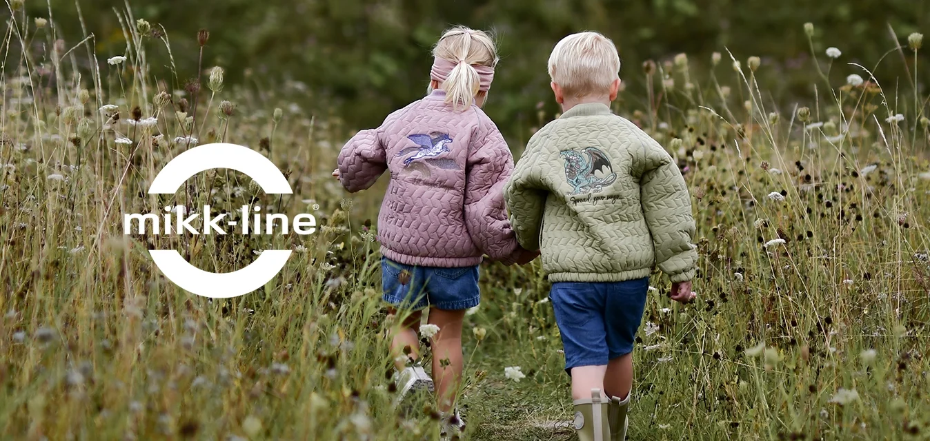 Due bambini mano nella mano che camminano su un sentiero stretto attraverso erba alta e fiori selvatici verso colline verdi sotto un cielo nuvoloso.