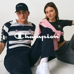 Un uomo e una donna sono seduti per terra contro una parete bianca, entrambi indossano abbigliamento casual Champion e mantengono pose rilassate.