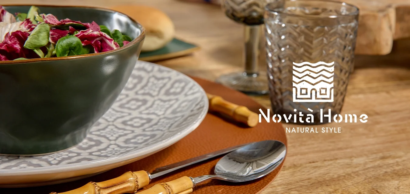 Ciotola di ceramica verde con insalata, piatto bianco decorato, posate con manico in bambù, bicchiere di vetro e sottopiatto marrone texture su tavolo di legno.