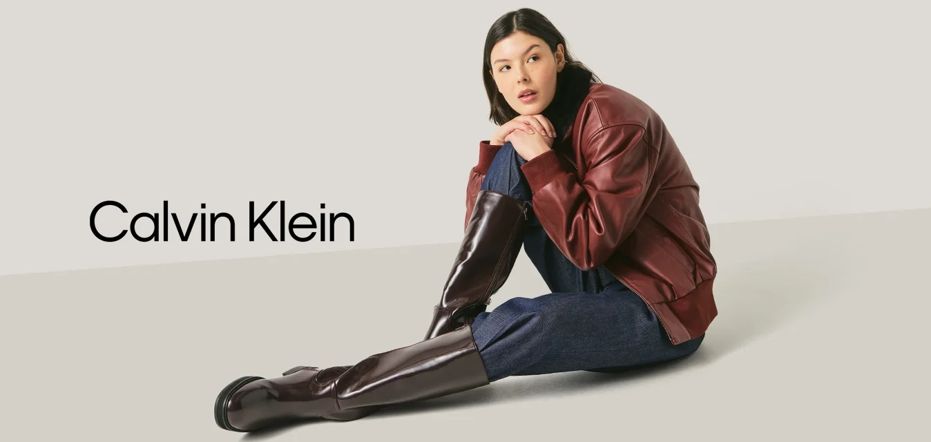 Giovane donna seduta per terra, indossa una giacca di pelle bordeaux, pantaloni blu scuro e alti stivali marrone scuro su uno sfondo chiaro e uniforme.