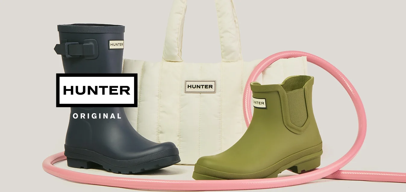 Stivale alto impermeabile blu navy, stivaletto corto verde oliva alla caviglia, borsa a tracolla bianca trapuntata, tutti con il logo Hunter, disposti insieme a un tubo rosa arrotolato.