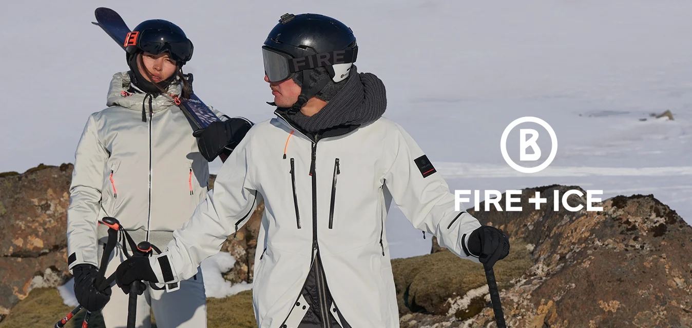 Sciatori in tute impermeabili bianche con dettagli neri si trovano sulla neve, tenendo i bastoncini da sci. Sullo sfondo si vedono terreni rocciosi e un cielo blu limpido.