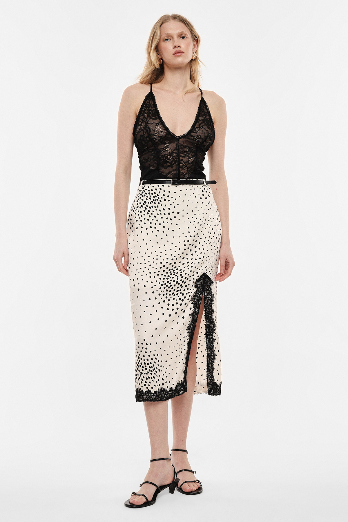 Woman modelling a black lace camisole with a cream midi skirt patterned with black polka dots and a side slit, black strappy sandals on white background