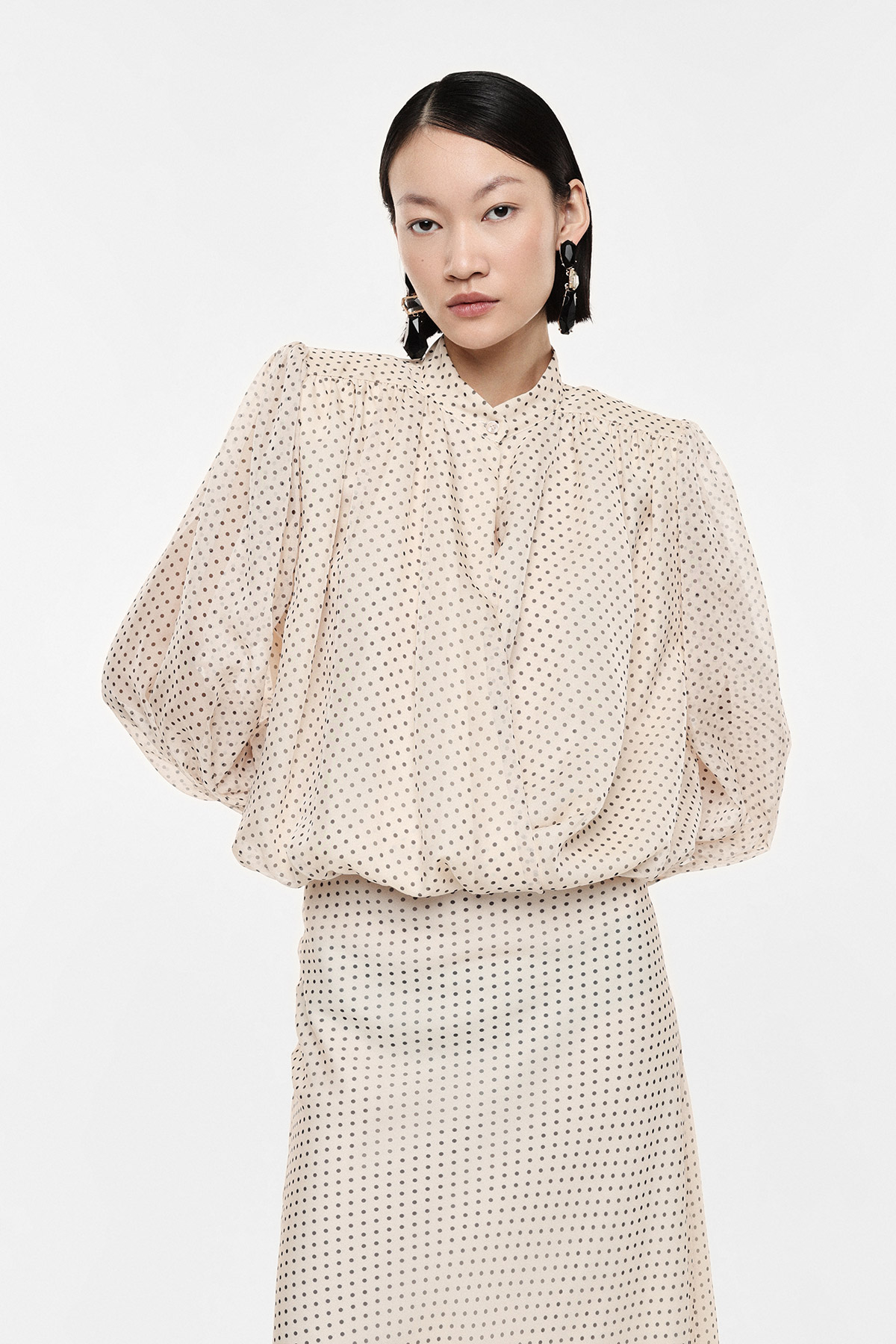 Woman modelling cream polka-dot blouse with voluminous sleeves and matching skirt, black tassel earrings, white background
