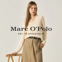 Donna con maglione color crema, gonna midi beige, cintura nera e mocassini neri in piedi accanto a un supporto pieghevole in legno su uno sfondo semplice.