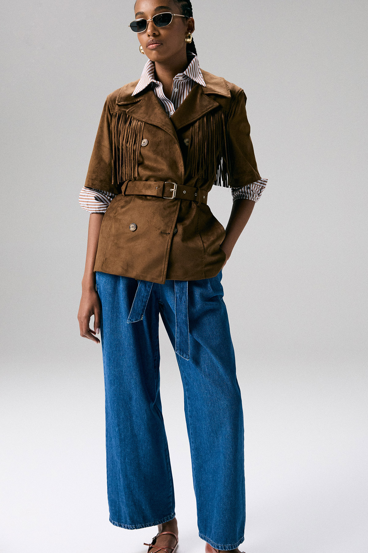 Model wearing brown suede fringed belted jacket, striped shirt, wide-leg blue jeans and sunglasses