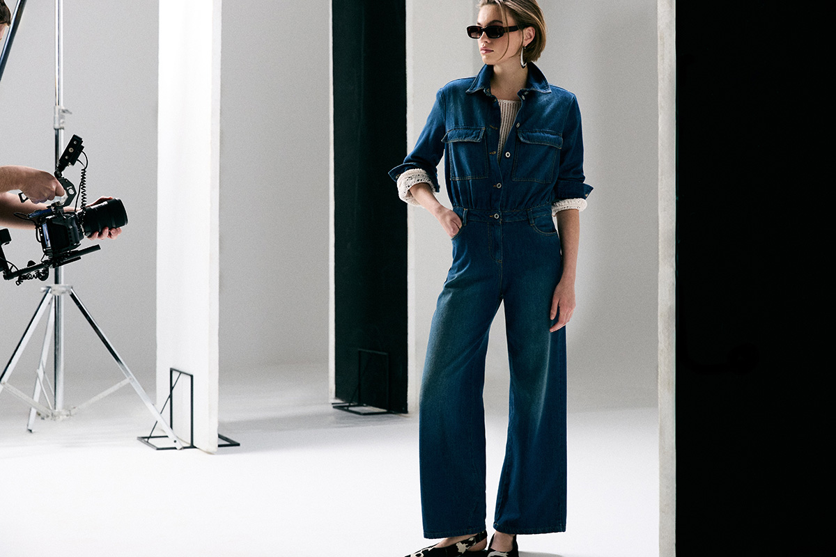 Full-length studio photo of a woman wearing a blue denim jumpsuit and sunglasses during a fashion photo shoot