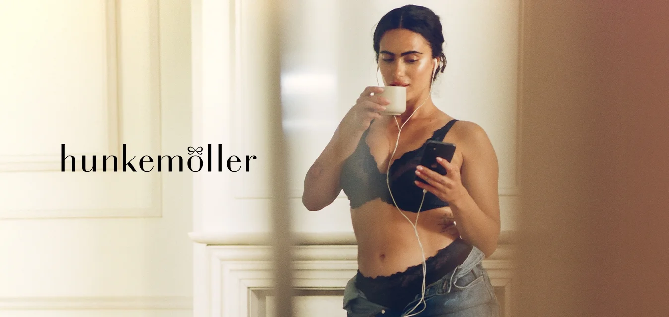 Bralette in pizzo nero con bordi smerlati, jeans in denim a vita alta e auricolari. Tiene una tazza bianca con una mano e uno smartphone con l'altra.