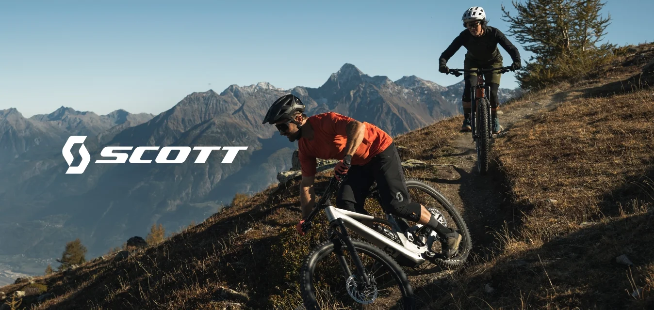 Due mountain biker con caschi sfrecciano giù per un sentiero ripido su una collina erbosa secca, con montagne alpine sullo sfondo.