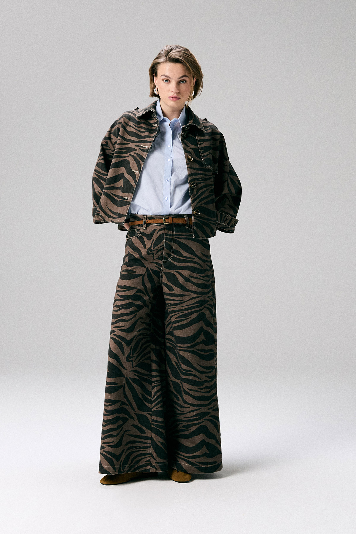 Woman wearing brown-and-black zebra-print jacket and wide-leg trousers with a light blue shirt and brown belt, standing