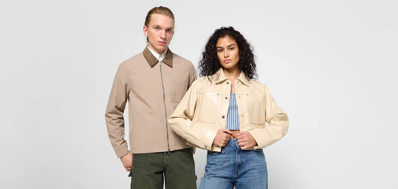 Giacca beige con zip e collo marrone, abbinata a una giacca in pelle crema corta sopra una maglietta a righe blu e jeans blu.