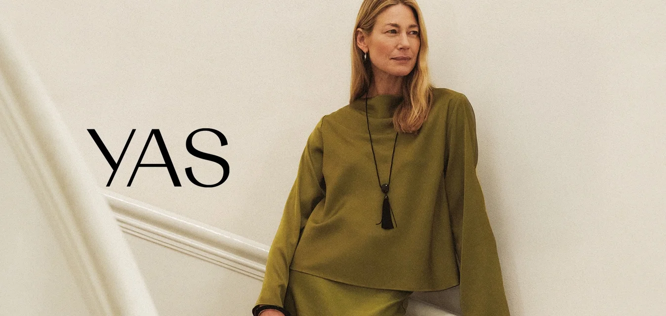 Donna con indosso un top a maniche lunghe verde oliva e gonna, che tiene in mano una borsa nera, in piedi su una scala in legno curva con pareti bianche e corrimano.