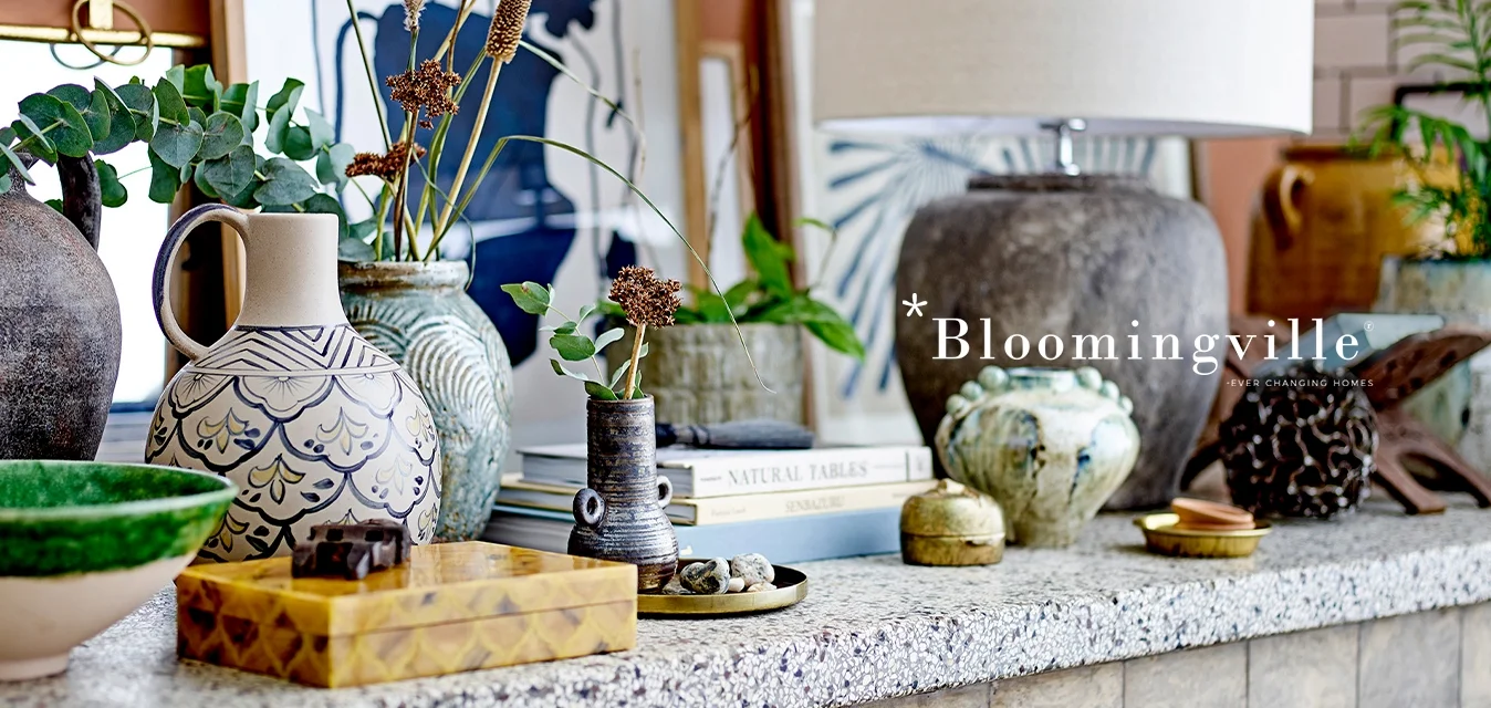 Vaso in ceramica con design floreale, ciotola verde, pezzi in stoneware texture, accenti in legno e libri decorativi su un tavolo maculato.