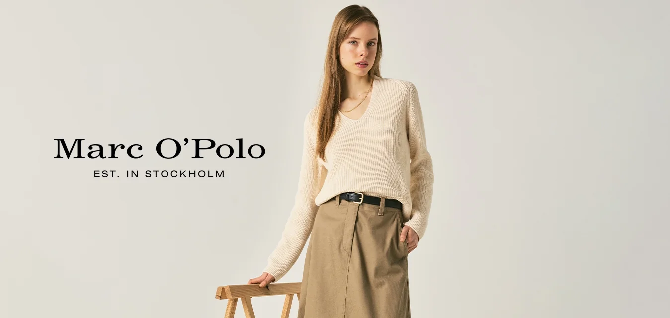 Donna con maglione color crema, gonna midi beige, cintura nera e mocassini neri in piedi accanto a un supporto pieghevole in legno su uno sfondo semplice.