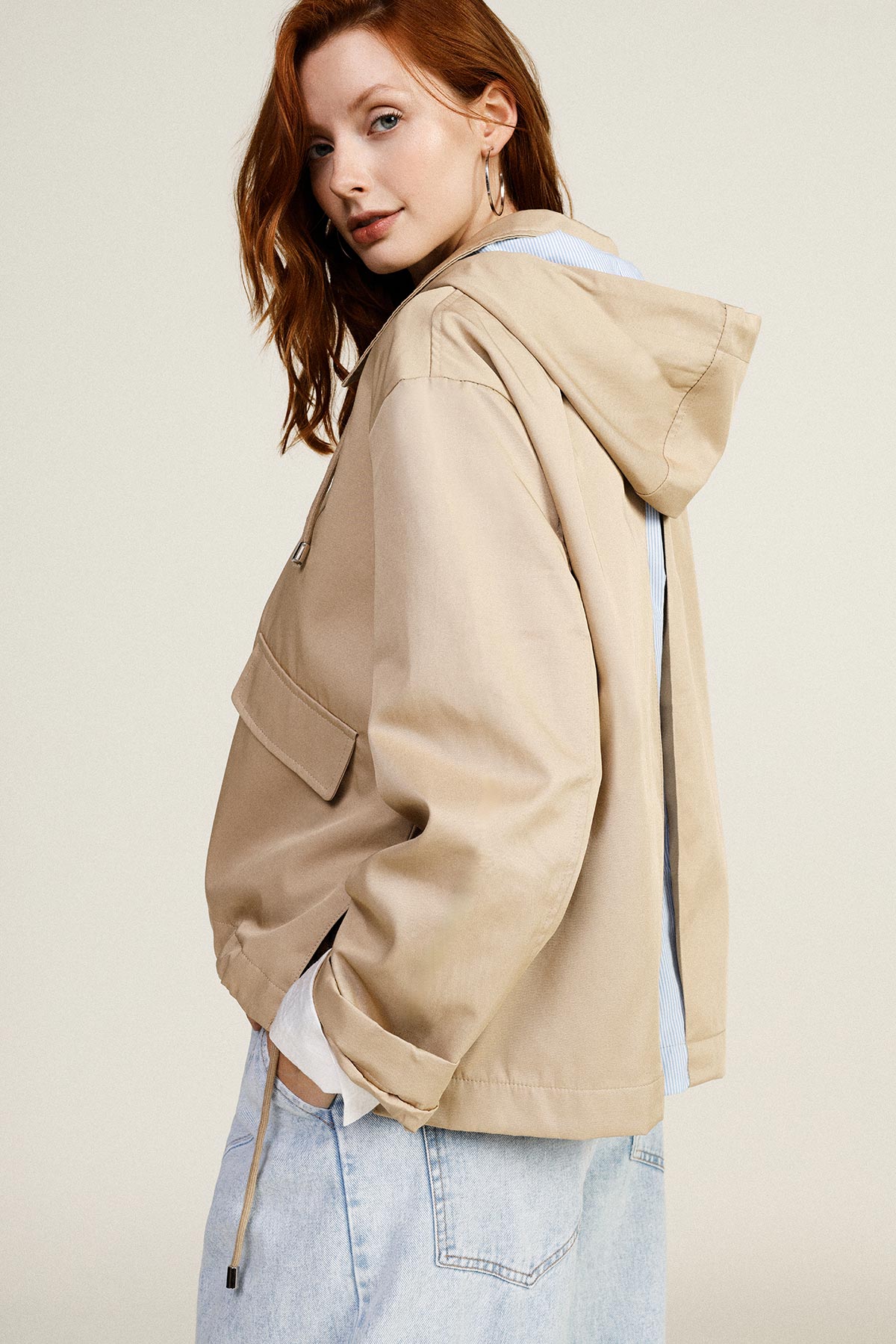 Woman in beige hooded jacket with side-slit back, light-blue shirt cuff and faded denim jeans in three-quarter pose