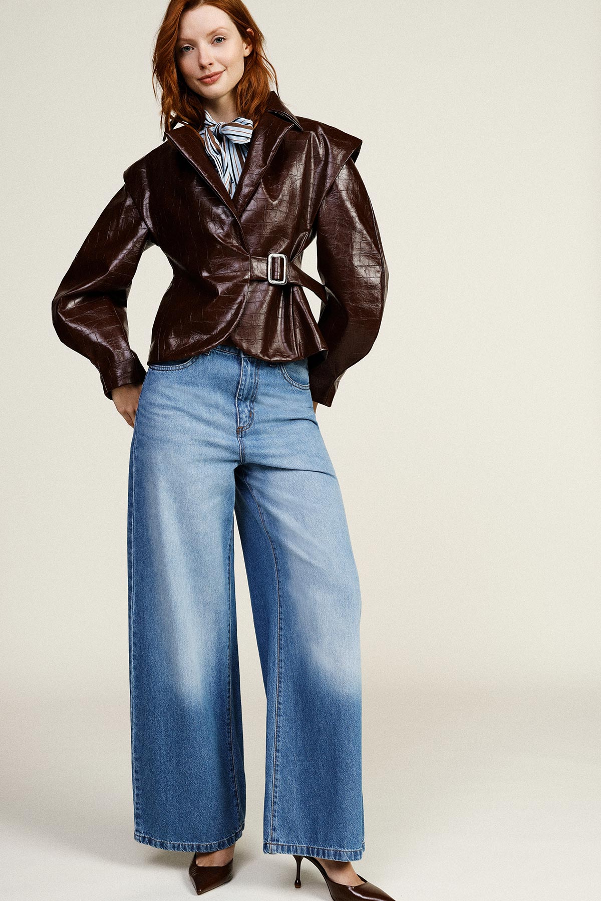 Model in brown belted leather jacket over a striped blouse, wide-leg light-blue jeans and brown heels on a neutral background