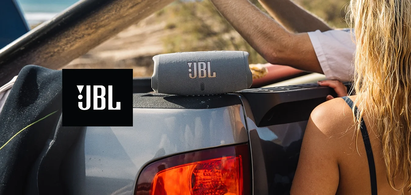 Altoparlante portatile JBL grigio su un cassone di un camion, affiancato da una muta nera e una maglietta a maniche lunghe bianca. Tavola da surf tenuta sopra la testa, ambiente da spiaggia.