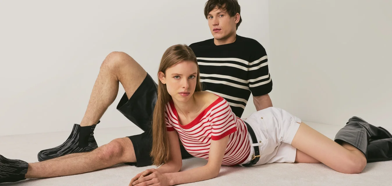 Giovane uomo e donna posano sul pavimento davanti a uno sfondo neutro, indossando magliette a righe, pantaloncini e stivali neri.