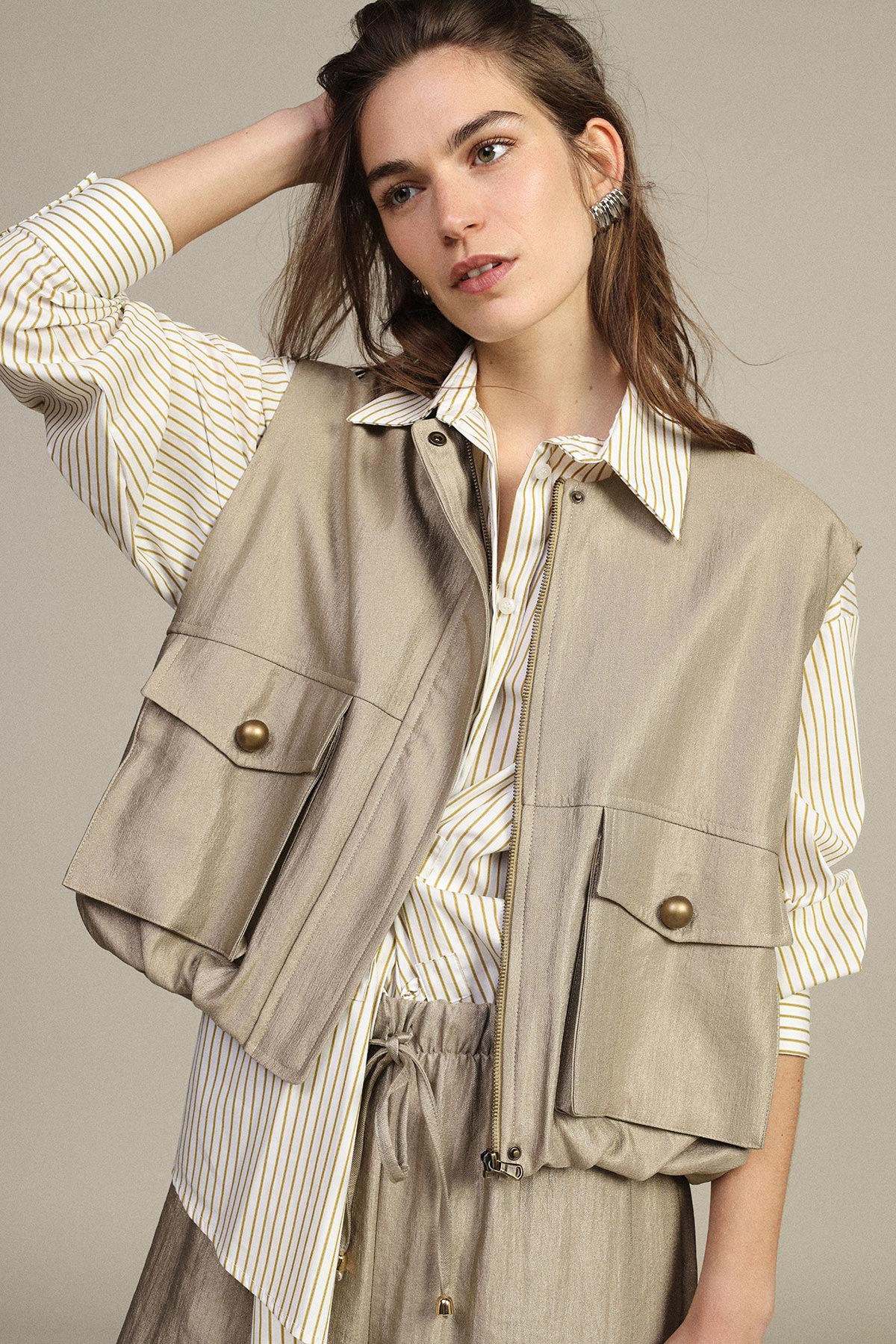 Young woman modelling beige utility vest with flap pockets over cream striped shirt and matching drawstring trousers against neutral background