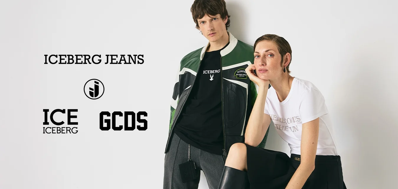 Due giovani adulti posano contro un muro bianco; uno in piedi con una giacca verde, l'altro seduto con una maglietta bianca e una gonna nera.