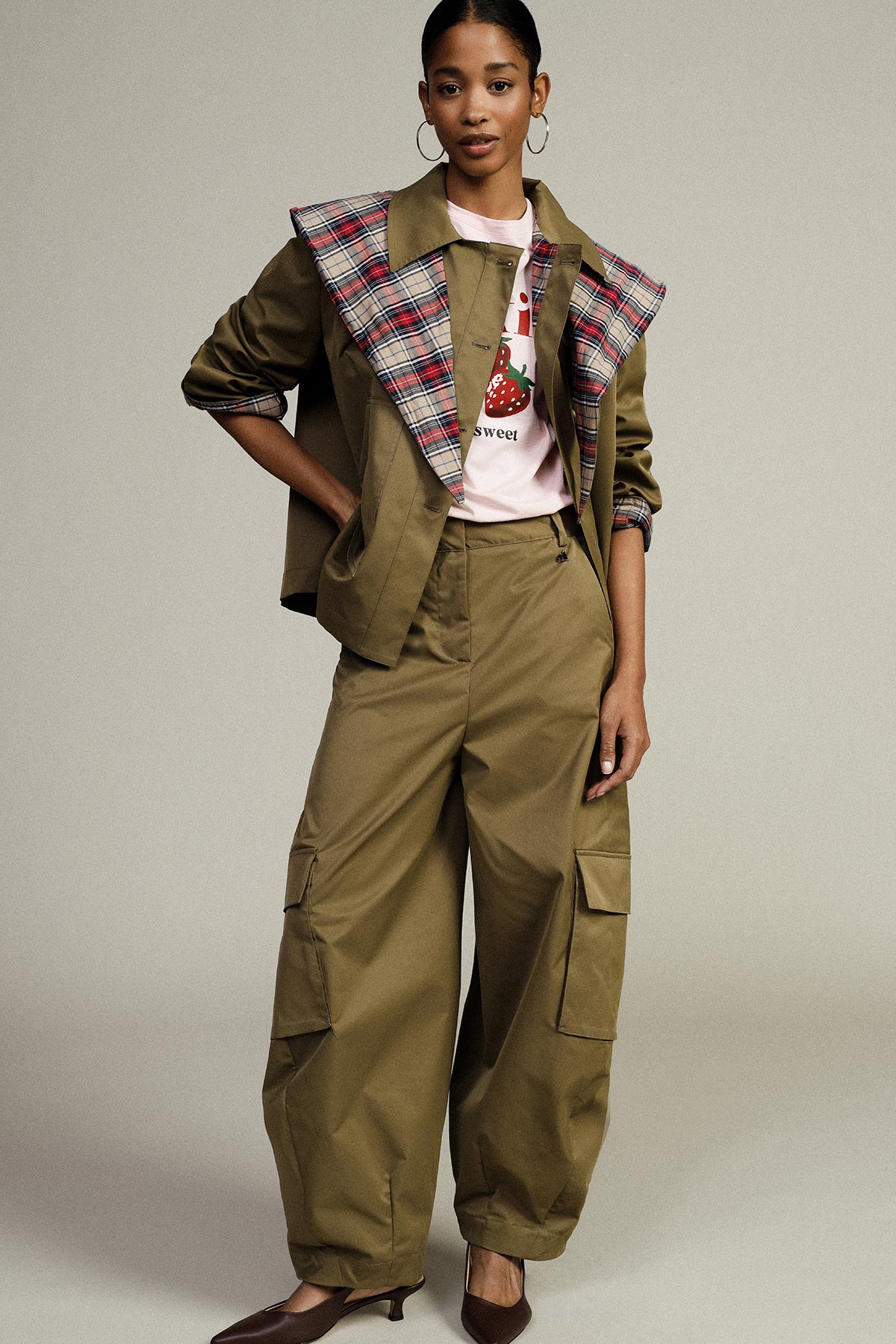 Woman modelling khaki cargo trousers and matching jacket with plaid collar, pink strawberry T-shirt, hoop earrings and heels in studio