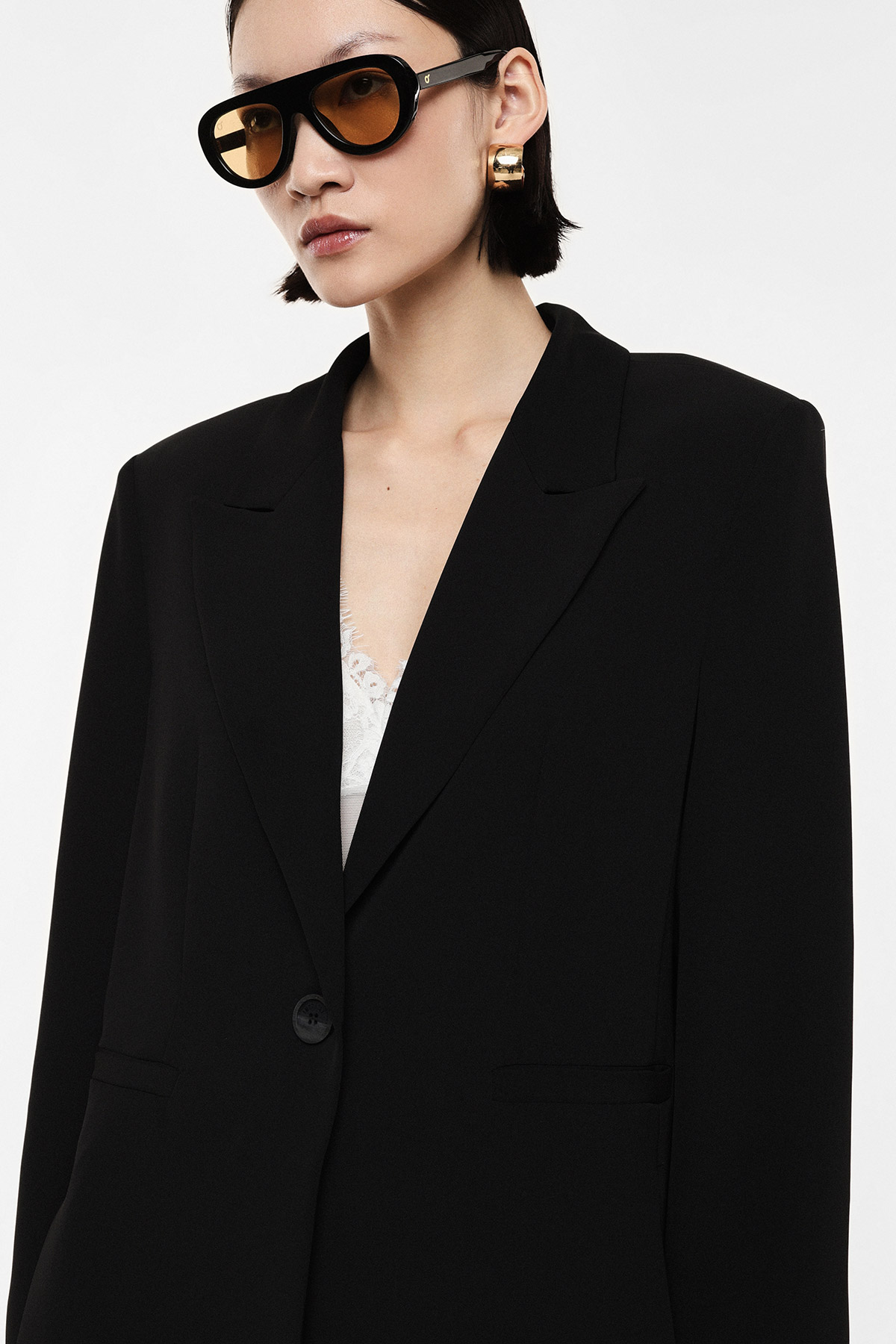 Woman wearing oversized black blazer and amber-lensed sunglasses, white lace top peeking and gold hoop earrings on white background
