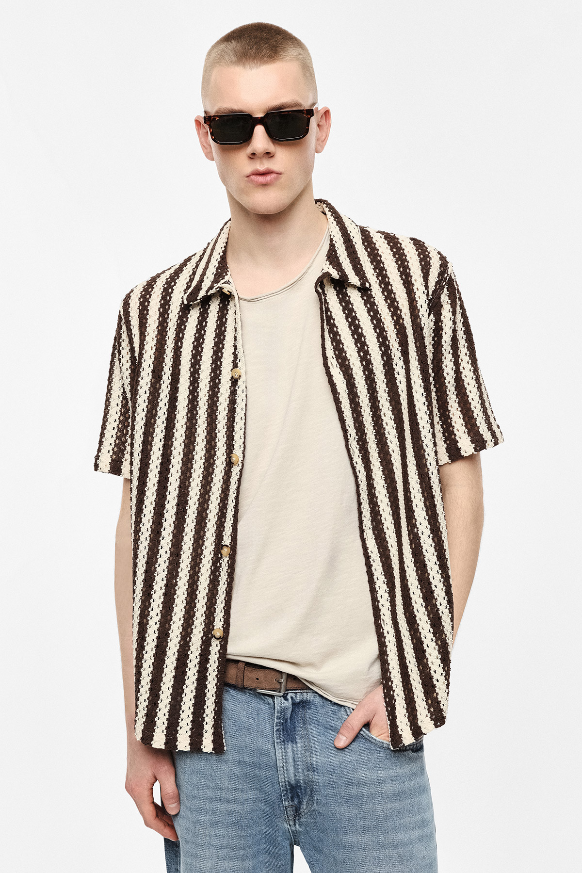 Young man wearing black sunglasses, brown-and-cream vertical striped short-sleeve shirt over beige T-shirt with blue jeans, hand in pocket