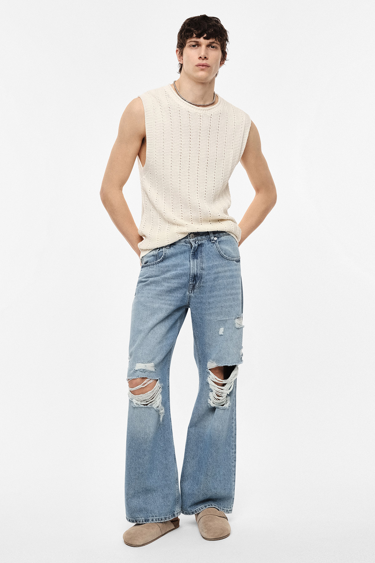 Male model in cream sleeveless knit top and distressed wide-leg blue jeans with beige slip-on shoes on white background