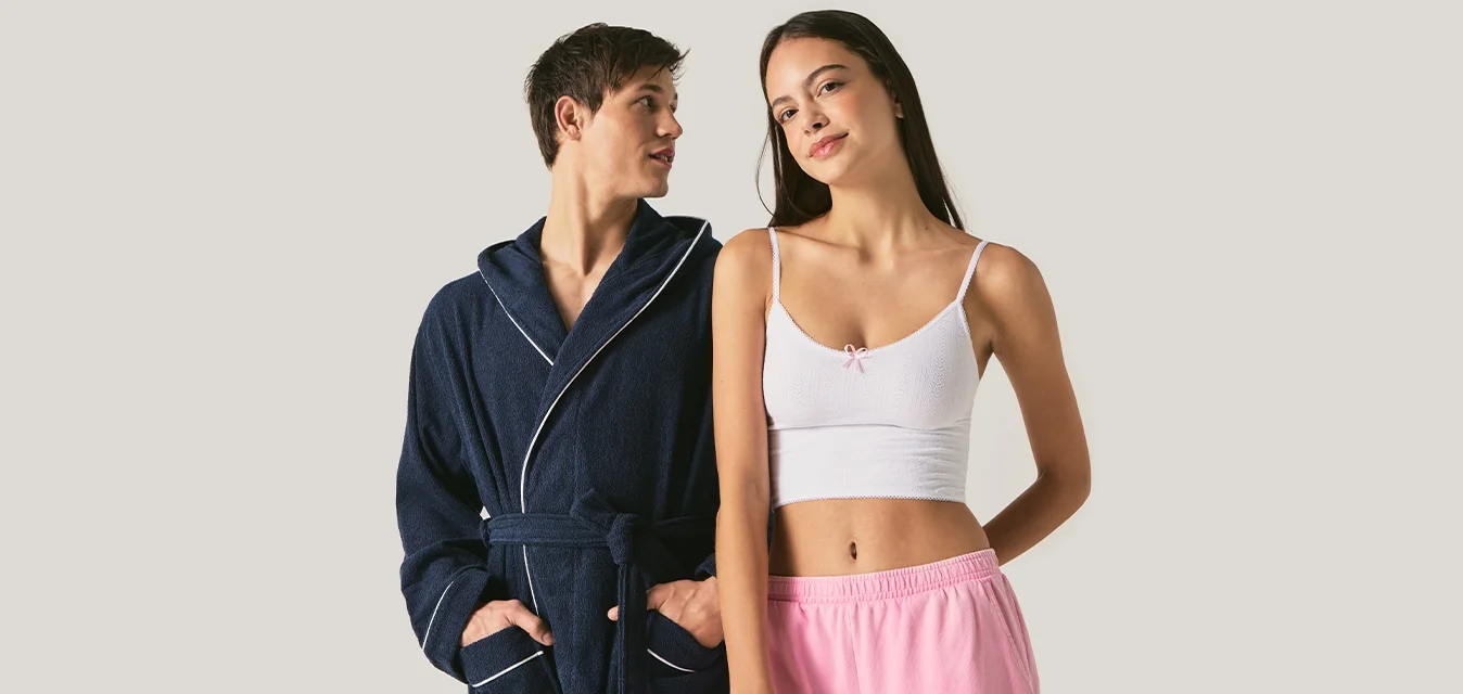 Giovane uomo con vestaglia blu navy e giovane donna con top corto bianco e pantaloni rosa stanno fianco a fianco contro uno sfondo chiaro e uniforme.