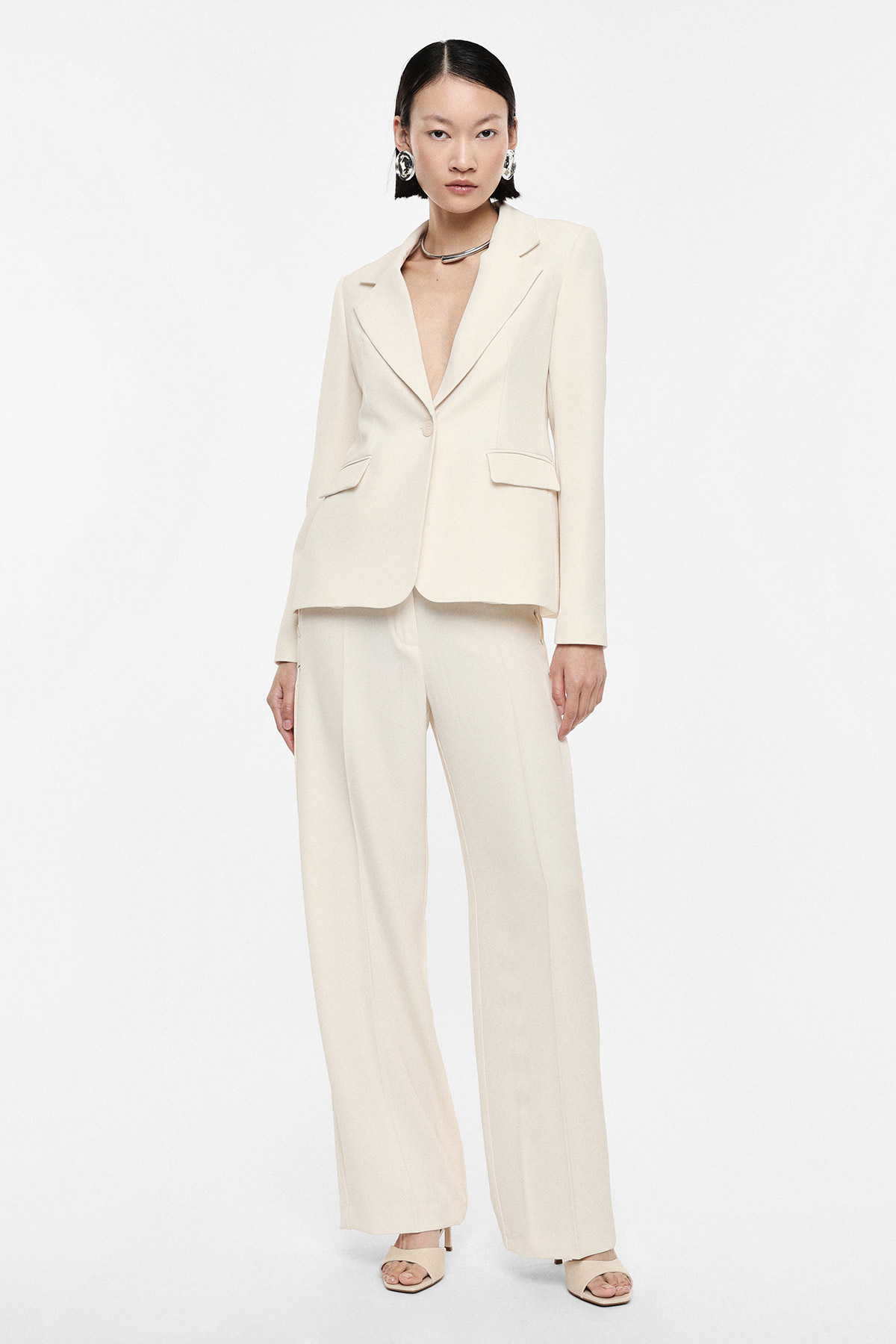 Woman modelling cream tailored blazer and wide-leg trousers with statement earrings and sandals on white background