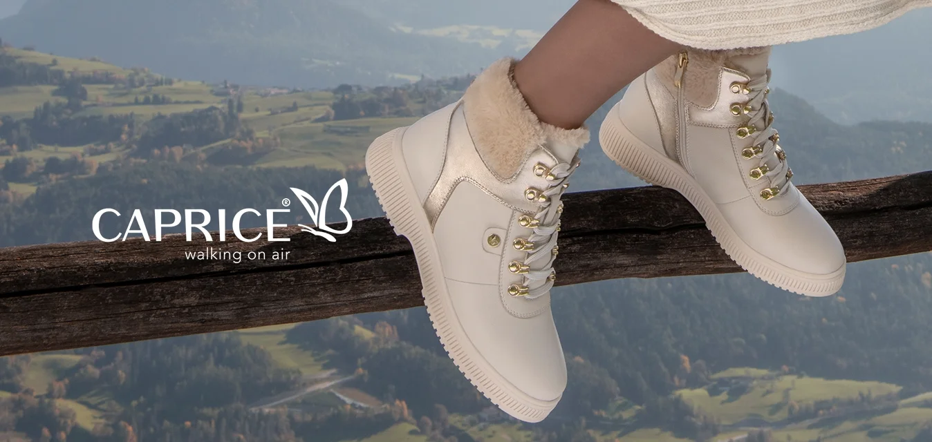 Cardigan beige lavorato a maglia, vestito bianco a coste, stivaletti bianchi alla caviglia con fodera in pelliccia e hardware dorato, seduta su un corrimano di legno con uno sfondo montano.