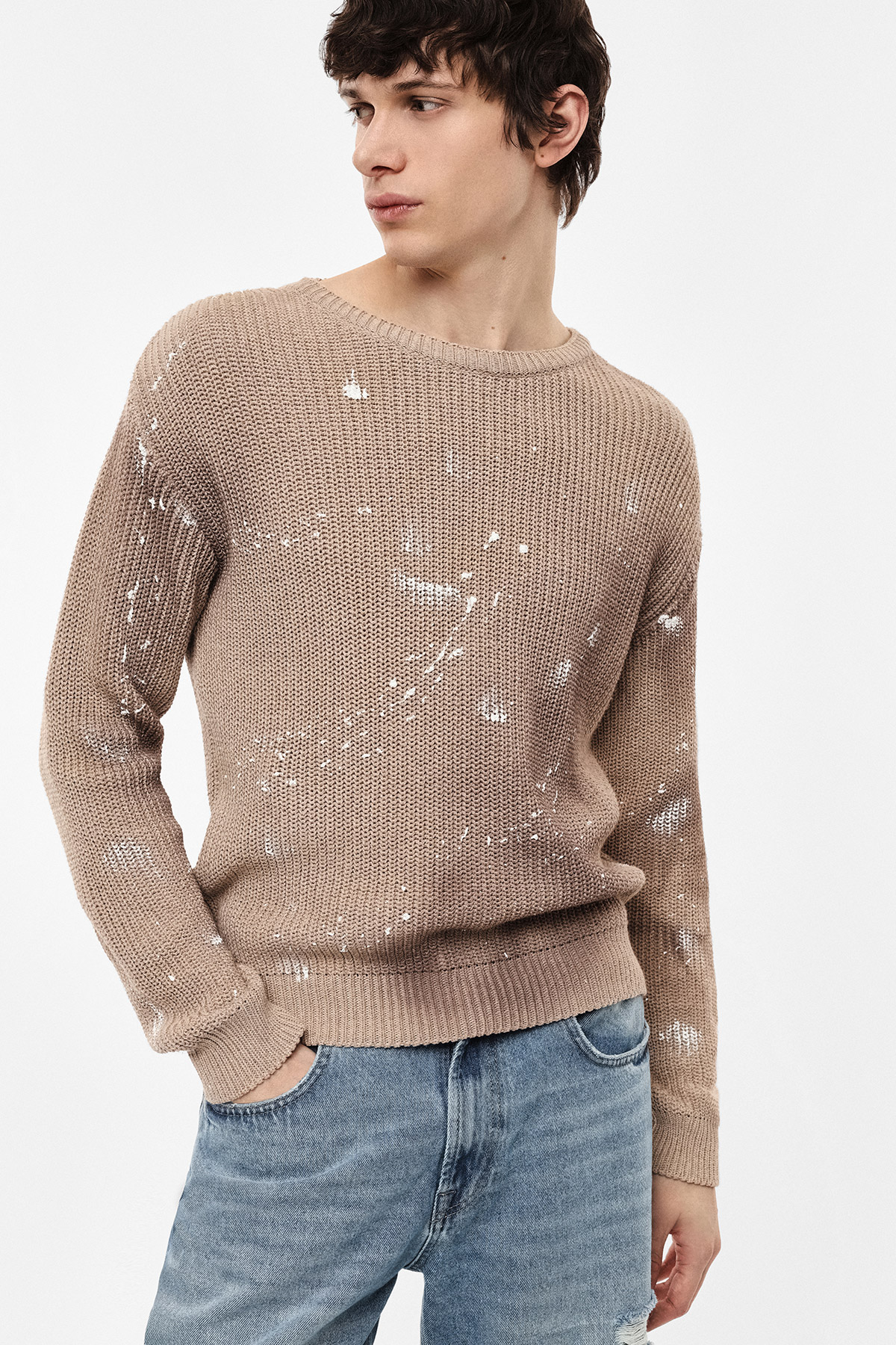 Young man modelling beige paint‑splattered knit sweater and blue jeans, hands in pockets, looking left on white background