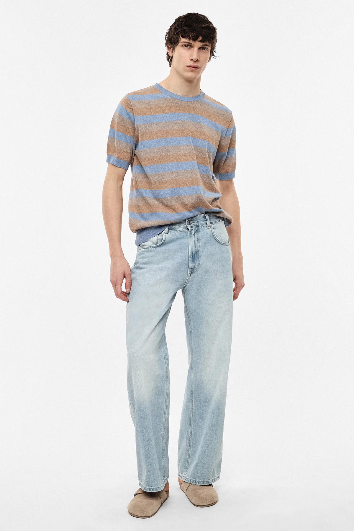 Young man wearing beige-and-blue striped short-sleeve jumper, light-wash wide-leg jeans and beige suede loafers on white background