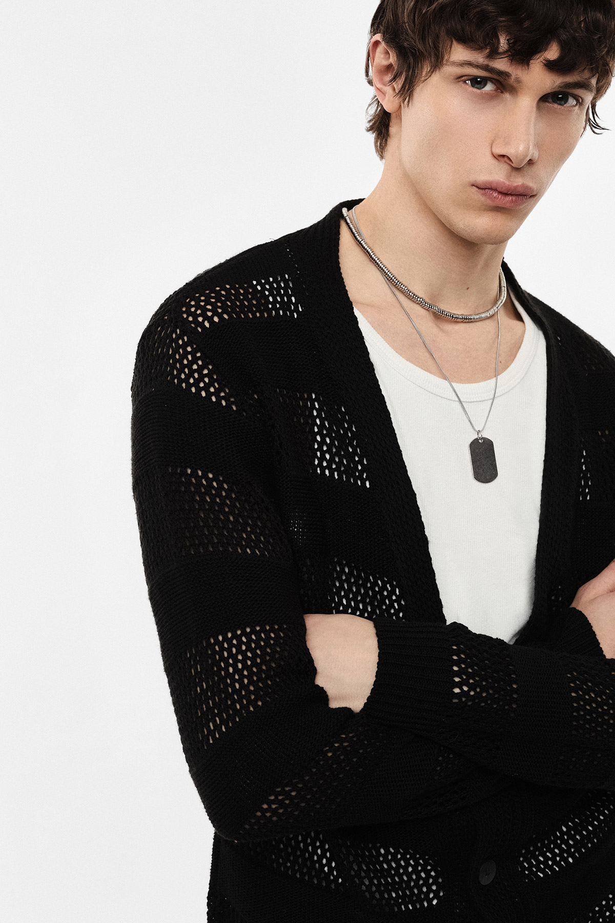 Young man wearing black open-knit cardigan over white ribbed vest, layered silver necklaces with dog tag, arms crossed