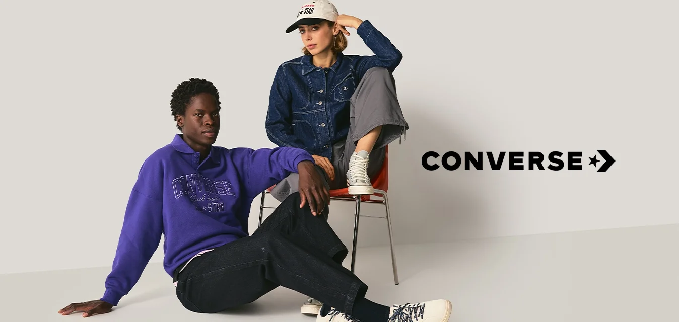 Due persone in abbigliamento casual; una seduta sul pavimento in un maglione viola, pantaloni neri e stivali bianchi; l'altra su sedia in giacca di jeans, pantaloni grigi e cappellino bianco.