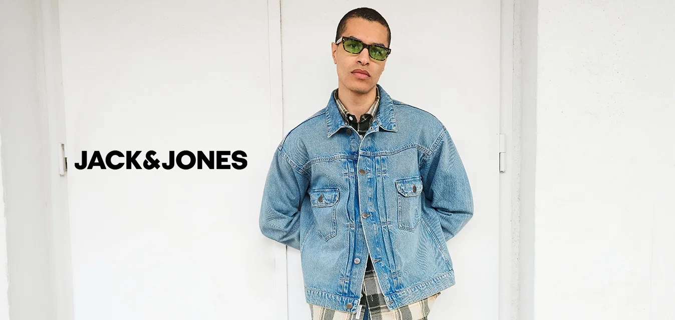 Uomo con occhiali da sole verdi, giacca di jeans chiara sopra camicia a quadri, jeans blu e scarpe nere, in piedi contro un muro bianco con le mani dietro la schiena.