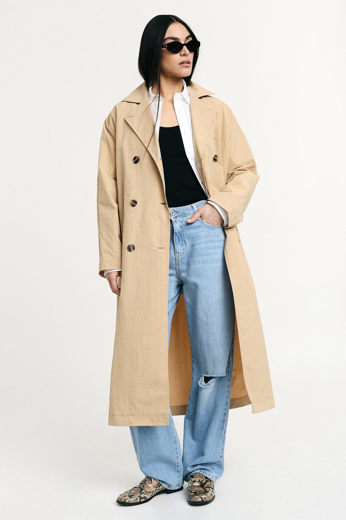 Woman model wearing beige trench coat with white shirt, black top, ripped light-blue jeans and snakeskin loafers, sunglasses