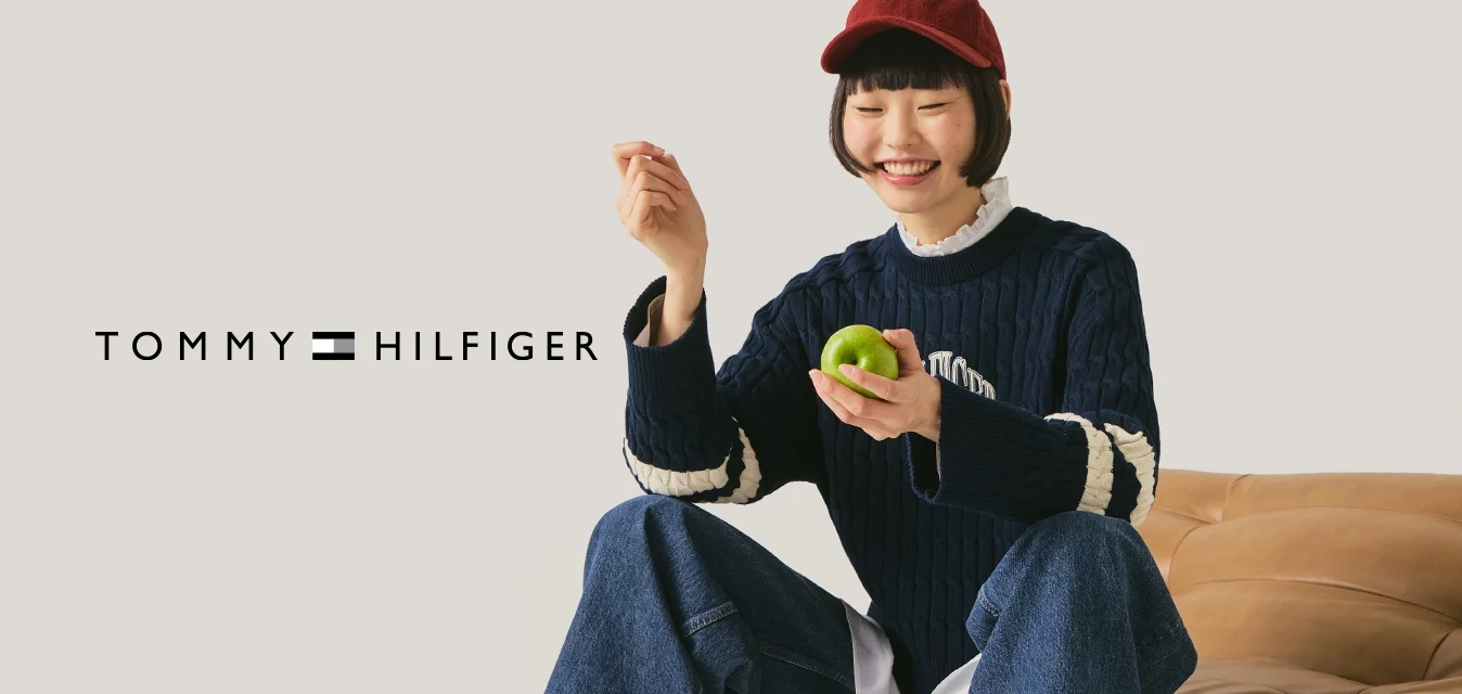 Giovane con un cappellino rosso, maglione blu navy e jeans seduto su un divano in pelle color cuoio, che tiene una mela verde sorridendo davanti a uno sfondo neutro.