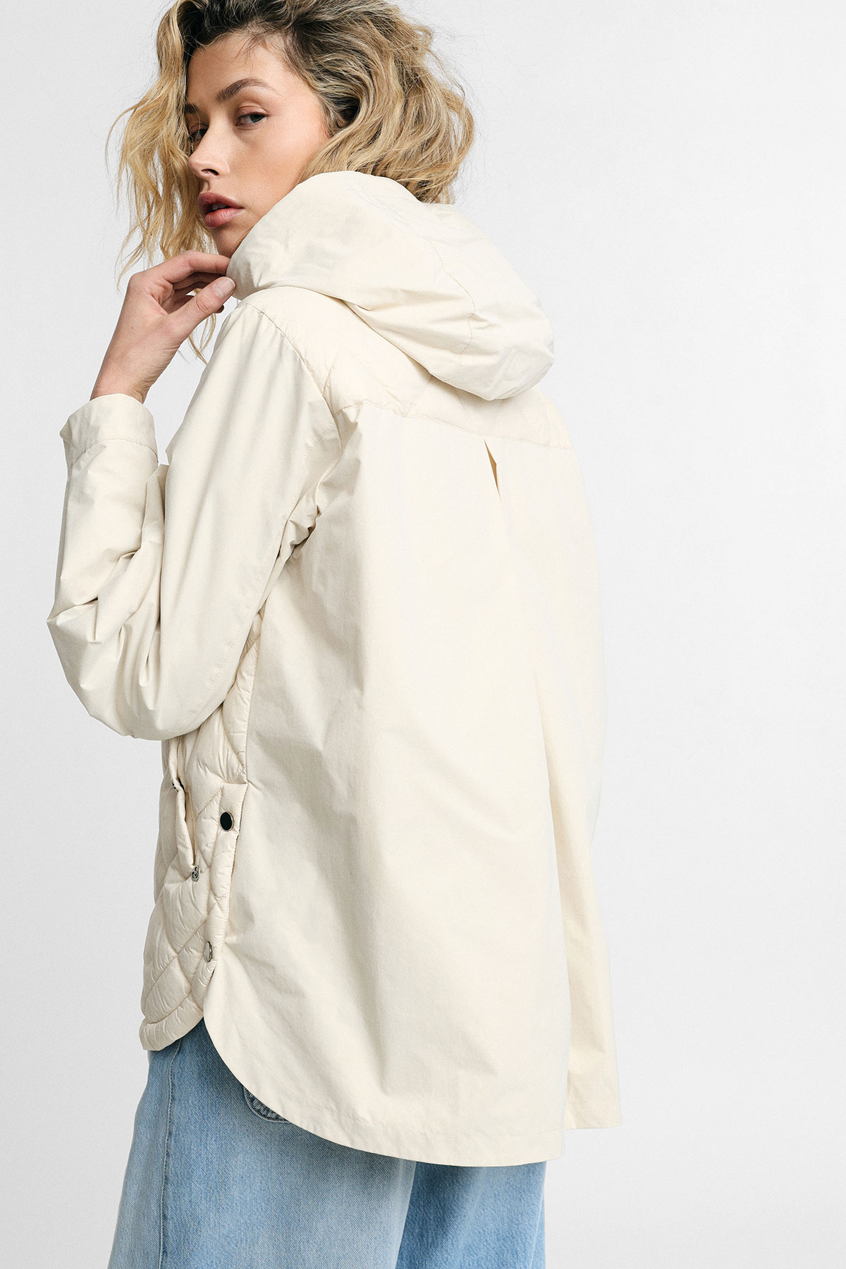 Woman wearing cream hooded rain jacket with quilted side panels, shown from back and side in light blue jeans