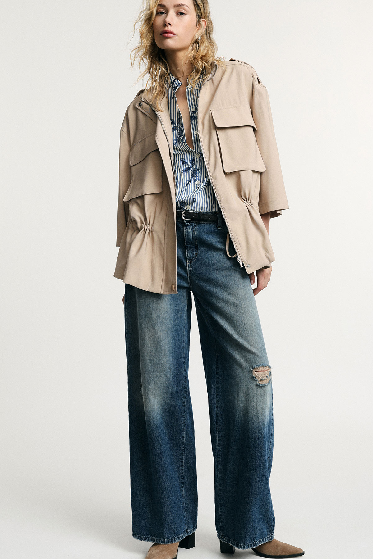 Woman wearing beige utility jacket over striped shirt with wide-leg distressed jeans and ankle boots, standing pose