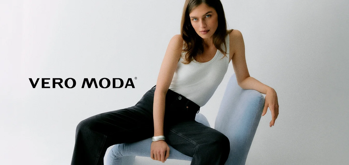 Donna con lunghi capelli castani seduta su una sedia moderna azzurro chiaro, indossa una canottiera bianca e jeans neri a gamba larga su sfondo bianco semplice.