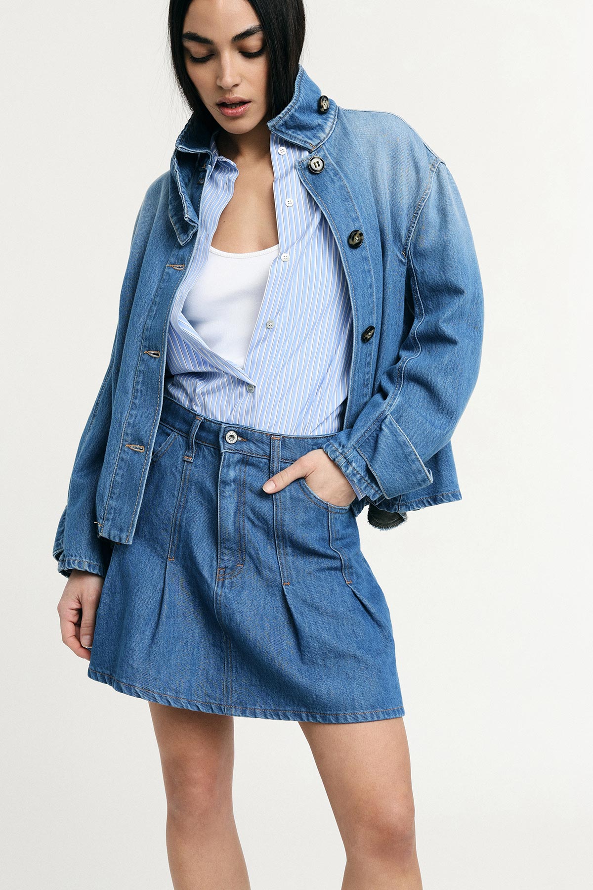 Woman wearing oversized denim jacket and high-waist denim skirt with blue striped shirt and white vest, hand in pocket