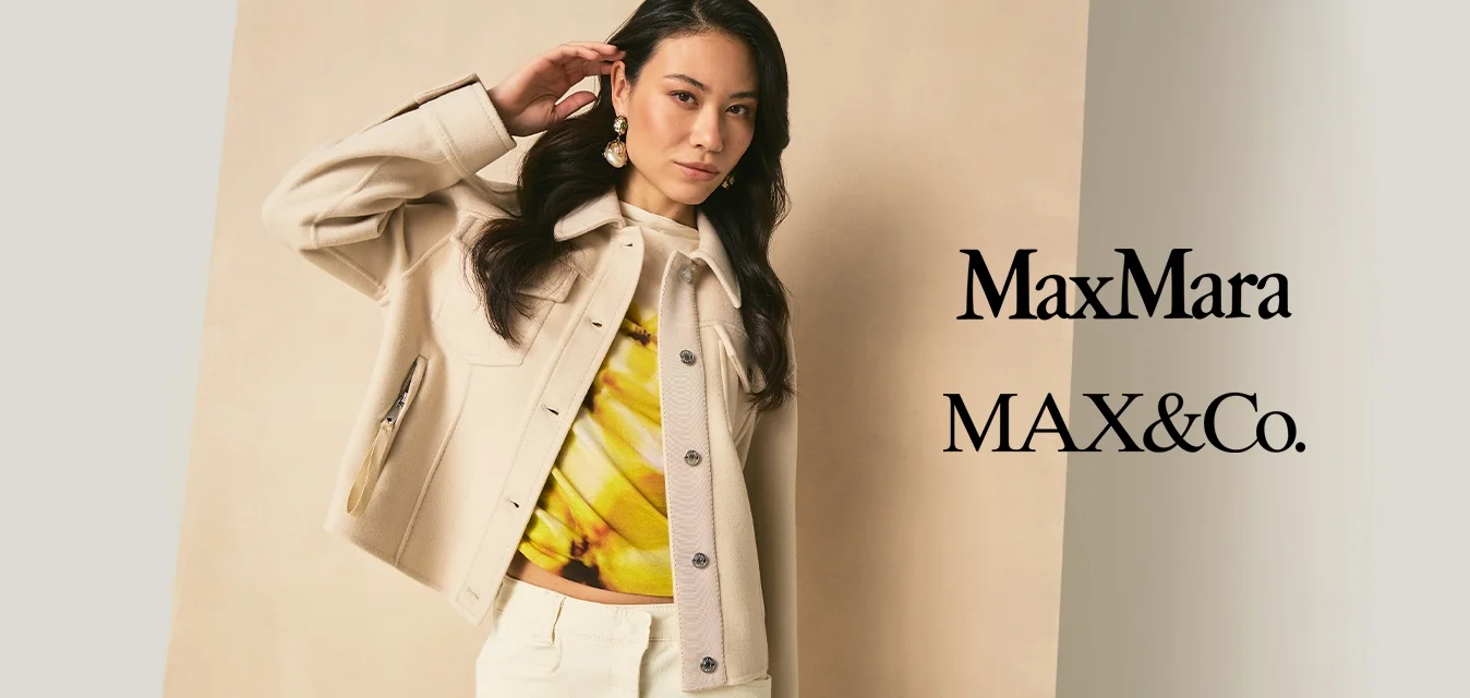 Donna con capelli lunghi e scuri che indossa una giacca beige, una maglia gialla con motivi e pantaloni chiari, in piedi davanti a uno sfondo neutro.