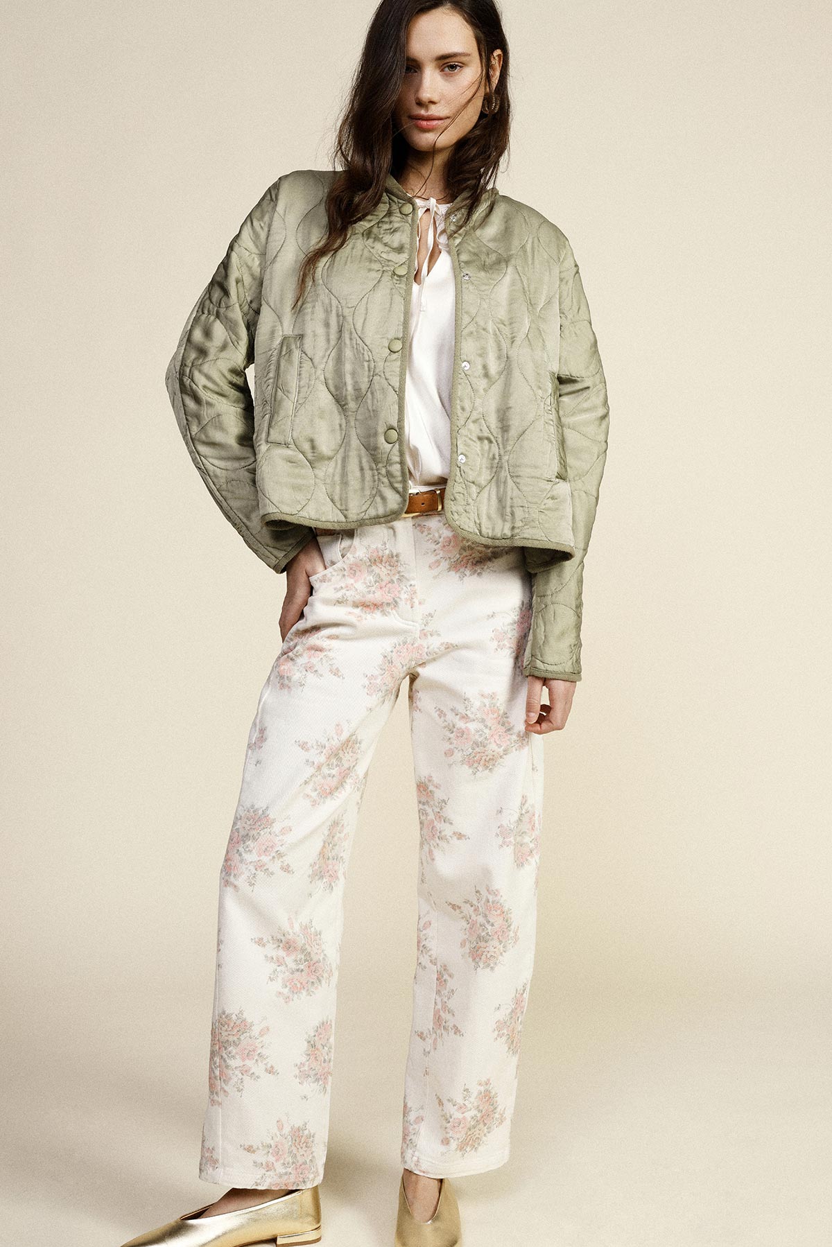 Model wearing quilted sage-green jacket over white blouse, high-waisted cream floral wide-leg trousers and gold flats against neutral background