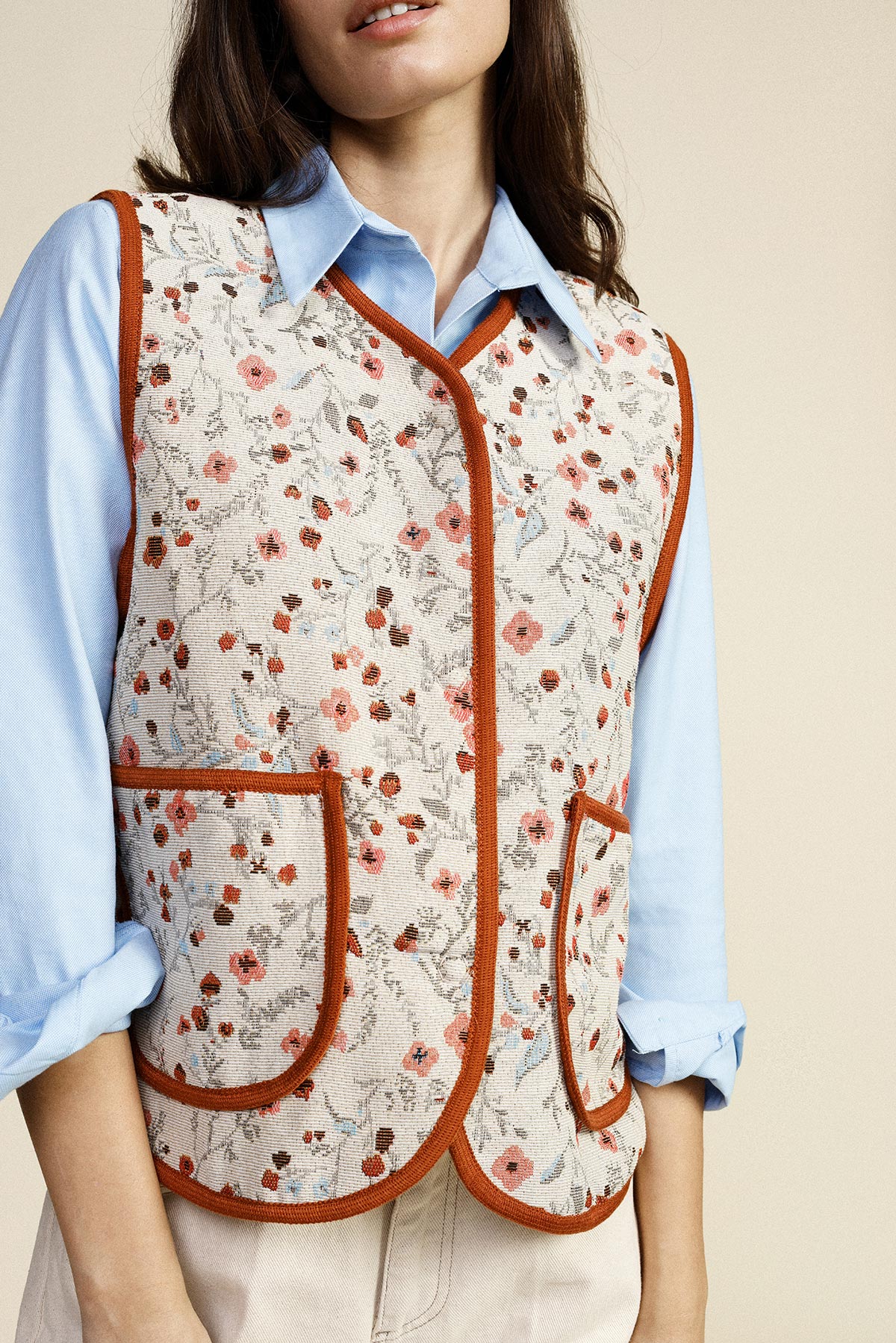 Woman wearing cream floral quilted waistcoat with rust trim over light-blue shirt, front pockets, casual spring outfit