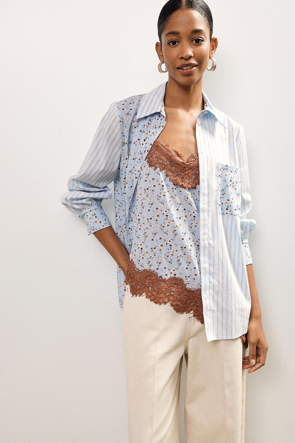 Model wearing light blue floral camisole with brown lace trim layered under striped button-up shirt and cream trousers
